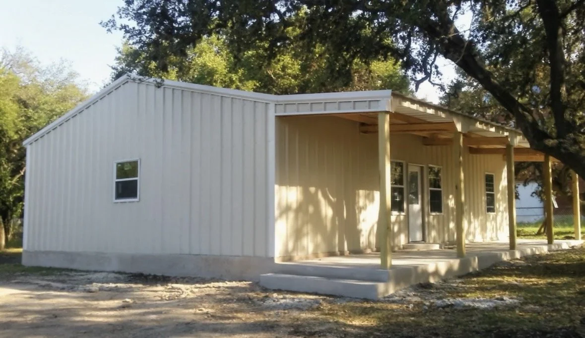 A newly built beige metal building with a covered porch supported by wooden posts, set in a rural area with trees nearby.