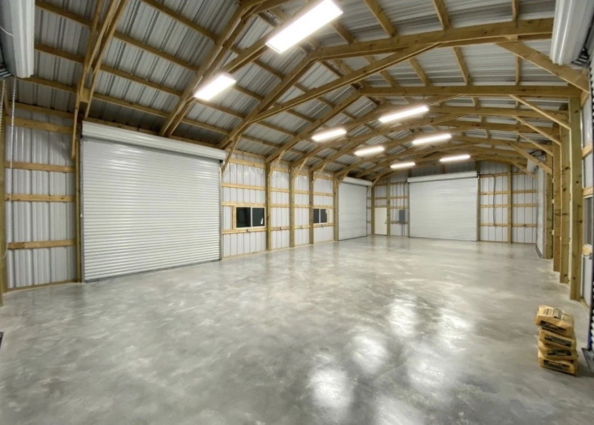 Empty metal warehouse with polished concrete floor, wooden framing, and fluorescent lights on the ceiling, with three large metal garage doors and small windows.