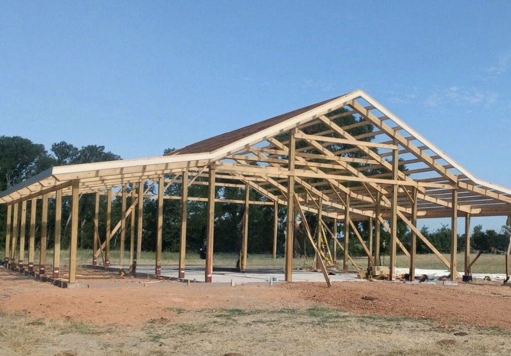 Construction site of a wooden building frame with a curved roof, set on a dirt lot with a clear blue sky