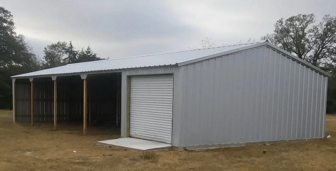 A large, metal storage shed with a partially enclosed area, a roll-up door, and a sloped roof, situated on a grassy area with trees in the background.