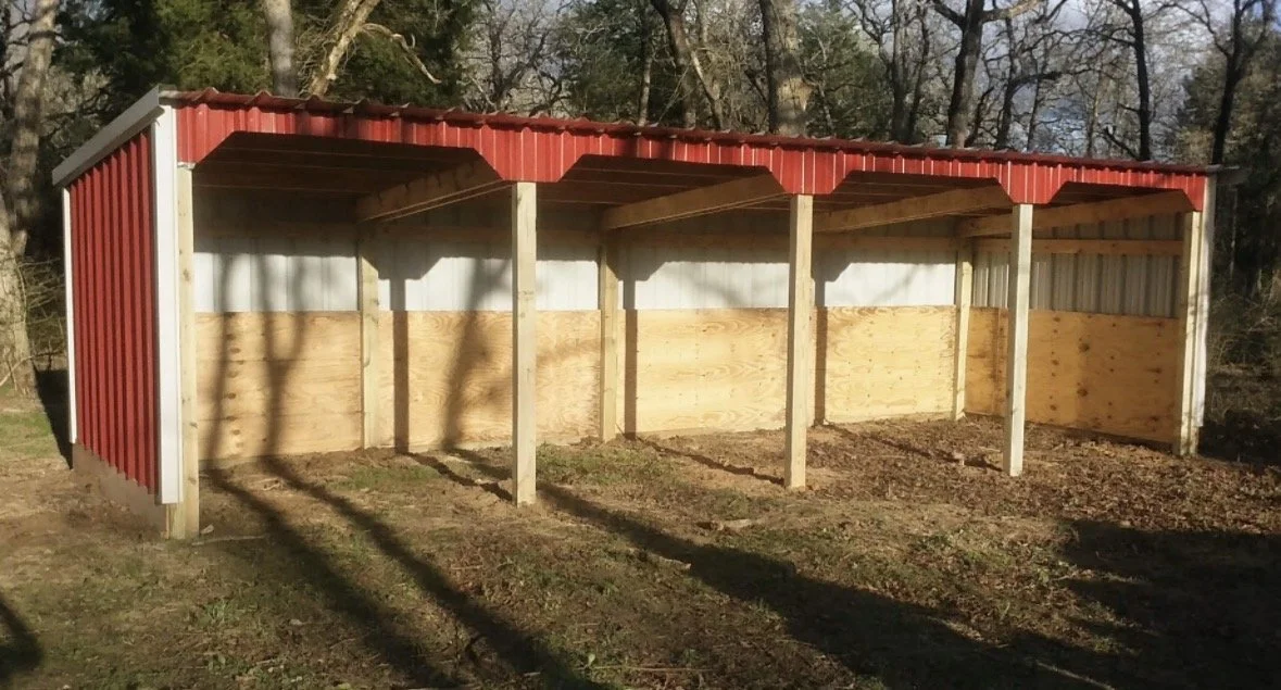 A partially built outdoor shed with a red corrugated metal roof, metal side panels, and wooden support beams and walls, situated on a grassy area with trees in the background.