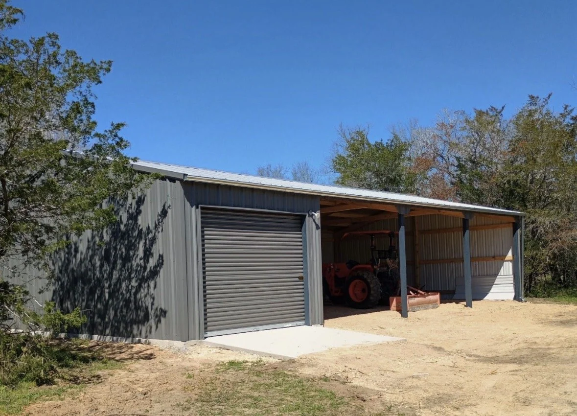 A gray metal storage shed with a closed roll-up door and an open side revealing a red tractor inside, set against a clear blue sky and surrounded by trees.