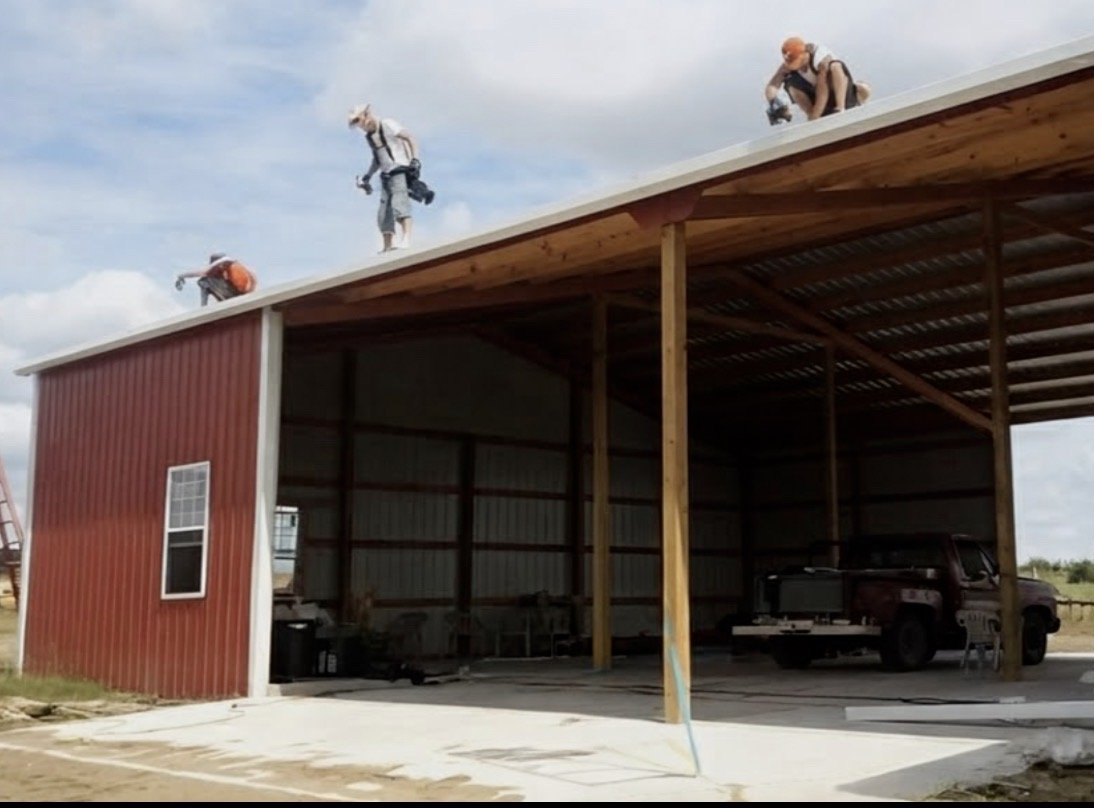 Three people installing a metal roof on a large open-sided shed, with one person on the roof and two others nearby.