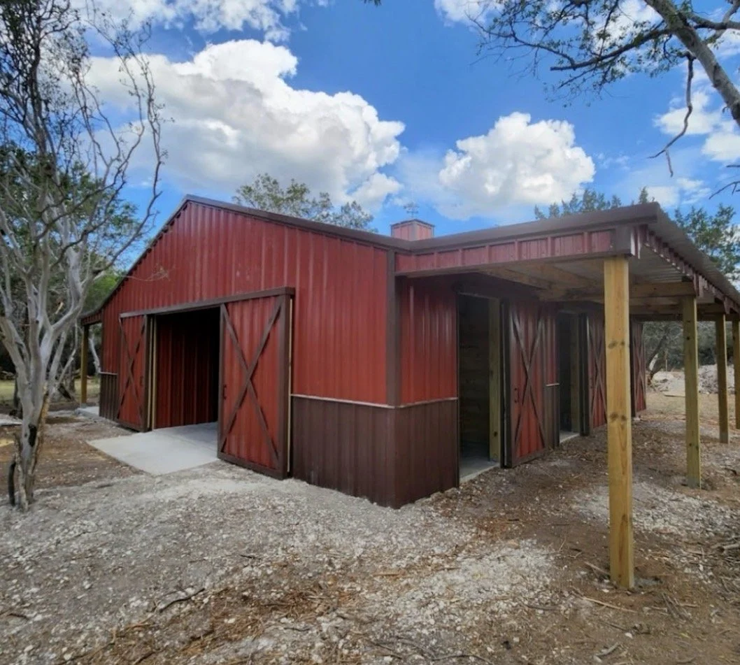 A red barn-style building under construction with open doors, surrounded by trees and a dirt ground during daytime.