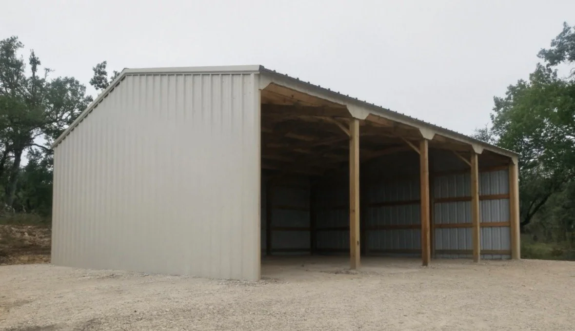 A metal storage shed with open front, metal walls, and wooden support beams inside, situated on a gravel lot surrounded by trees.
