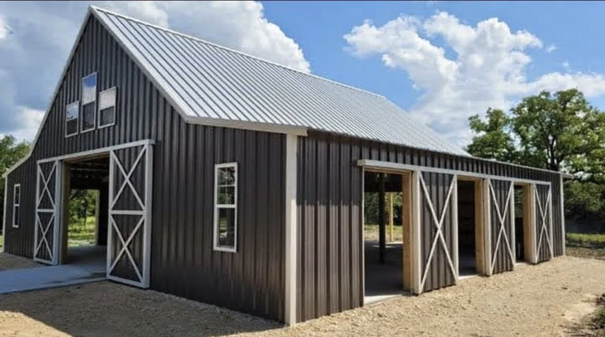 A black barn with white trim under construction, featuring a large sliding door, multiple smaller windows, and unfinished sections.