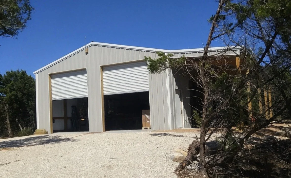 A metal building with two large garage doors, situated on a gravel lot, with a tree on the right and a clear blue sky in the background.