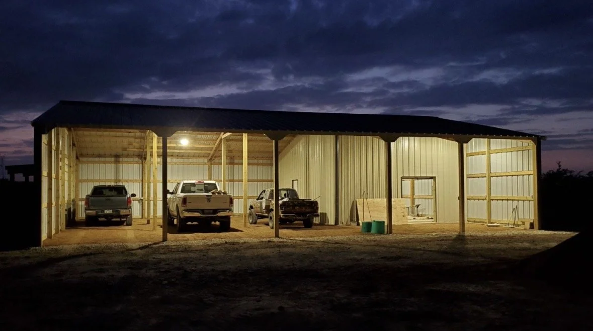 A metal carport with three vehicles parked inside during dusk, with a partially cloudy sky overhead.