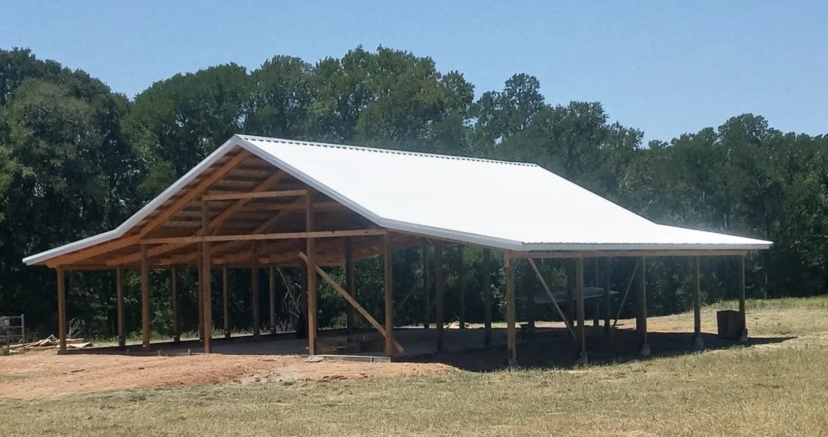 A wooden building under construction with a metal roof, surrounded by grass and trees.