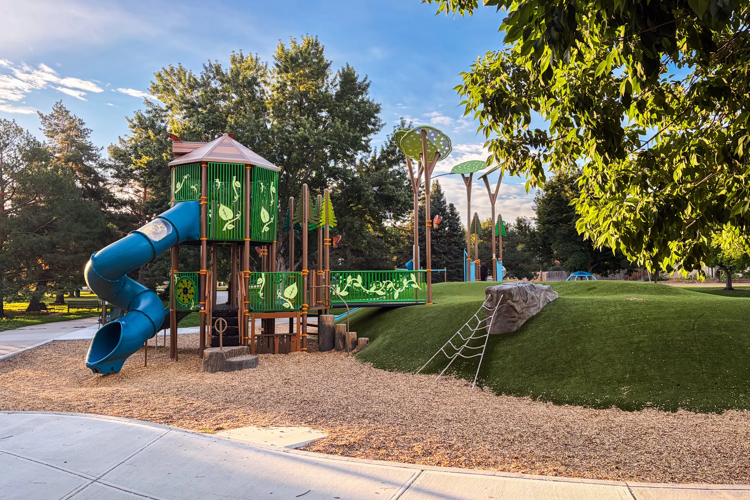 A playground with green and blue play structures, including a slide, surrounded by trees and a grassy area, with a sunny sky and some clouds.