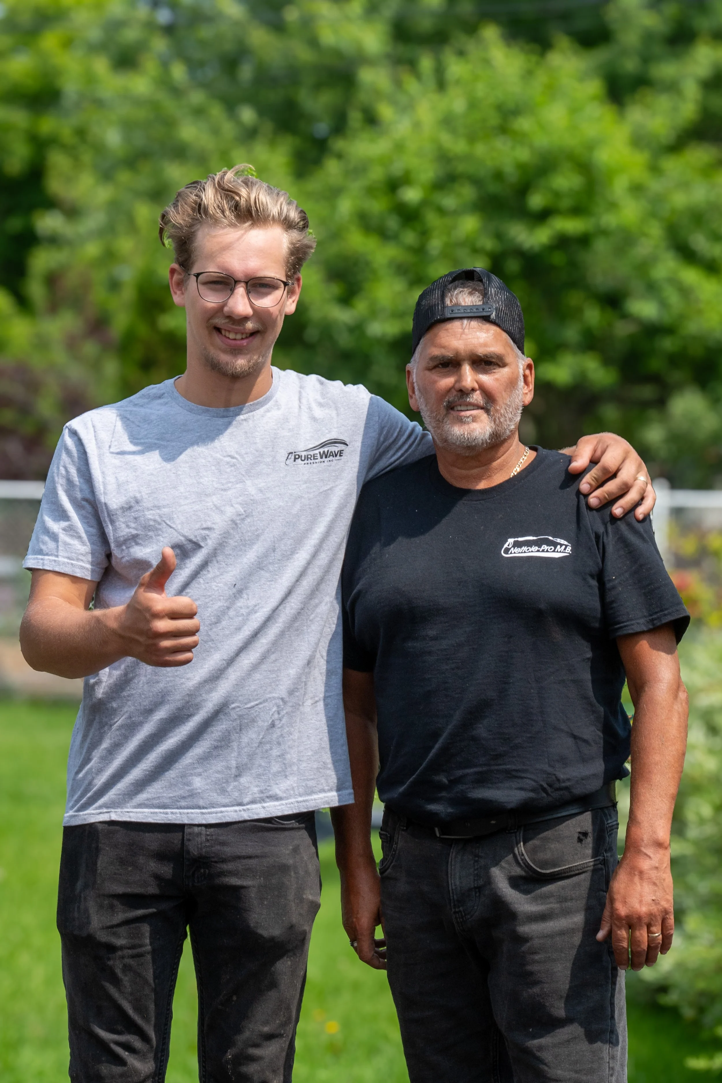 Two men stand together outdoors with green trees in the background, smiling and posing for the photo. The man on the left has light skin, blond hair, glasses, and is wearing a light gray t-shirt and black jeans, giving a thumbs-up. The man on the right has darker skin, gray hair, and is wearing a black cap, black t-shirt, and jeans, with his arm around the other man's shoulder.