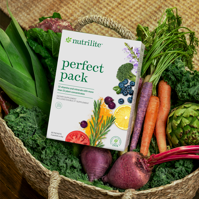 A basket filled with fresh vegetables including lettuce, carrots, purple carrots, beets, artichoke, and greens, with a Nutrilite Perfect Pack supplement box placed on top of the vegetables.
