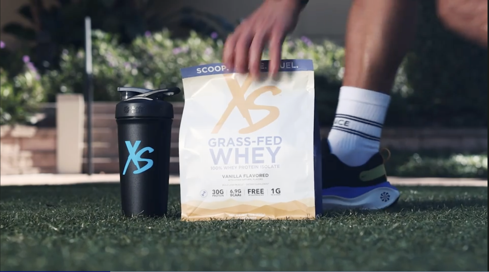 A black shaker bottle and a bag of X's Grass-Fed Whey protein powder on a grassy outdoor surface, with a person's hand reaching for the bag. The person's leg is visible, wearing a sock and athletic shoe.