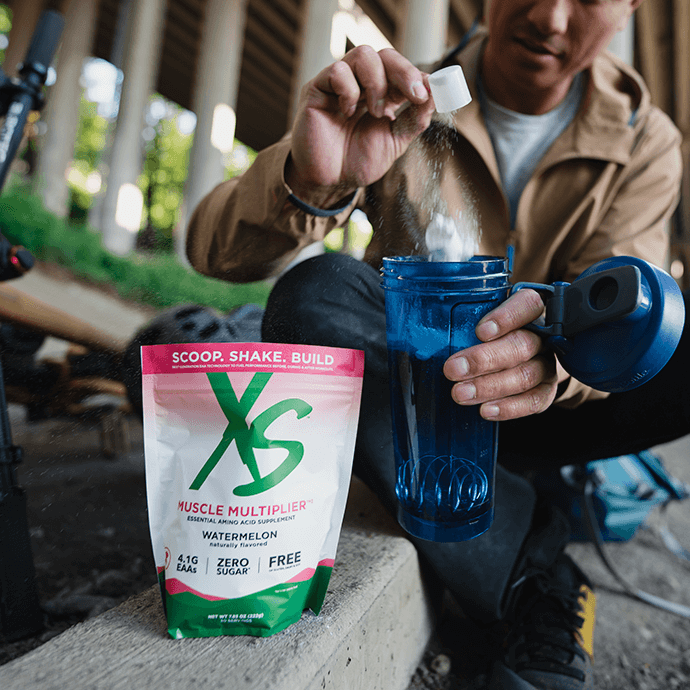 Person pouring powdered supplement into a blue water bottle outdoors with fitness gear nearby.