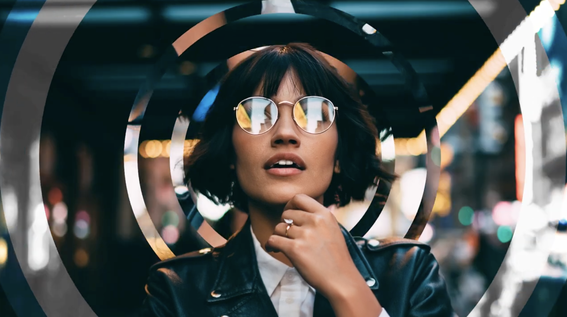 A woman with glasses and short dark hair posing with city lights in the background, framed within concentric circular shapes.
