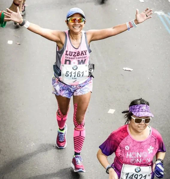 A woman in colorful running attire celebrating during a marathon, with her arms raised, wearing sunglasses, a cap, and a race bib with the number 51161. Another woman nearby is wearing a pink shirt, sunglasses, and a purple visor.