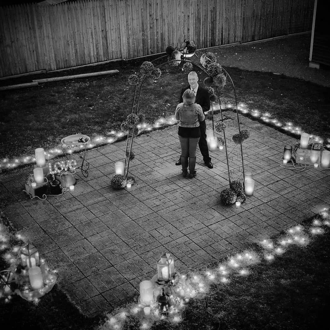 A couple sharing a kiss in a decorated outdoor space at night, with string lights, candles, and floral arrangements creating a romantic setting.