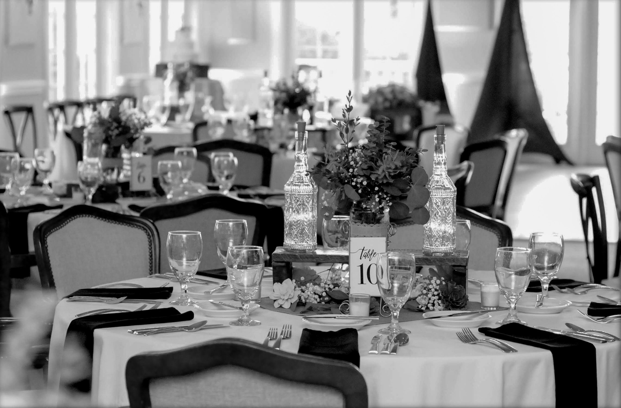 A banquet table decorated with floral centerpieces in glass bottles, surrounded by wine glasses, silverware, and black napkins, set for a formal event in a well-lit room with large windows.