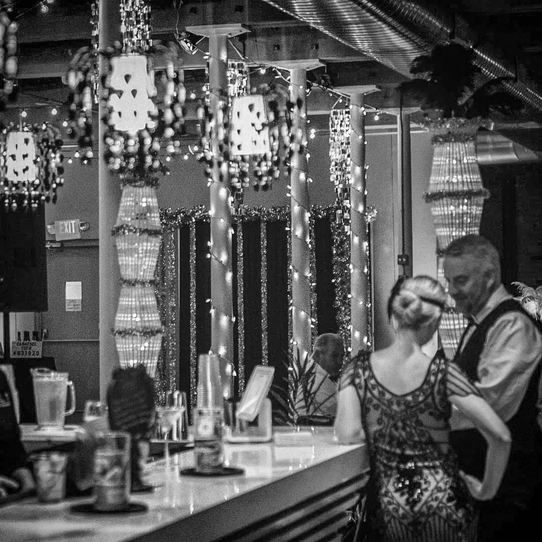A black and white photo of a lively bar or restaurant with hanging decorative lights and ornaments. In the foreground, a woman with her hair styled in an updo, wearing a patterned dress, is engaging with a man in a vest. In the background, a man sits at the bar with a drink. The setting has warm lighting and festive decorations.
