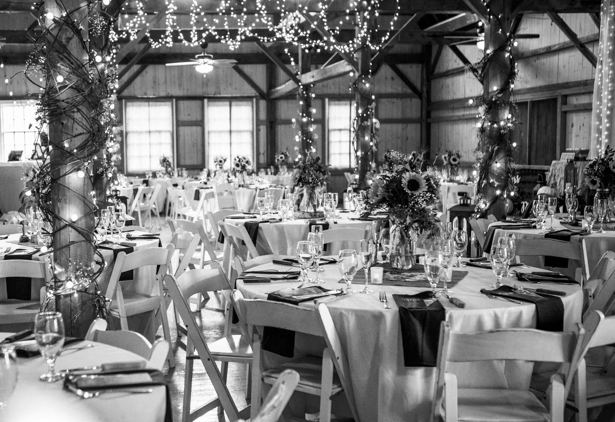 Decorated banquet hall with tables set for a celebration, surrounded by string lights, floral centerpieces, and wooden beams.