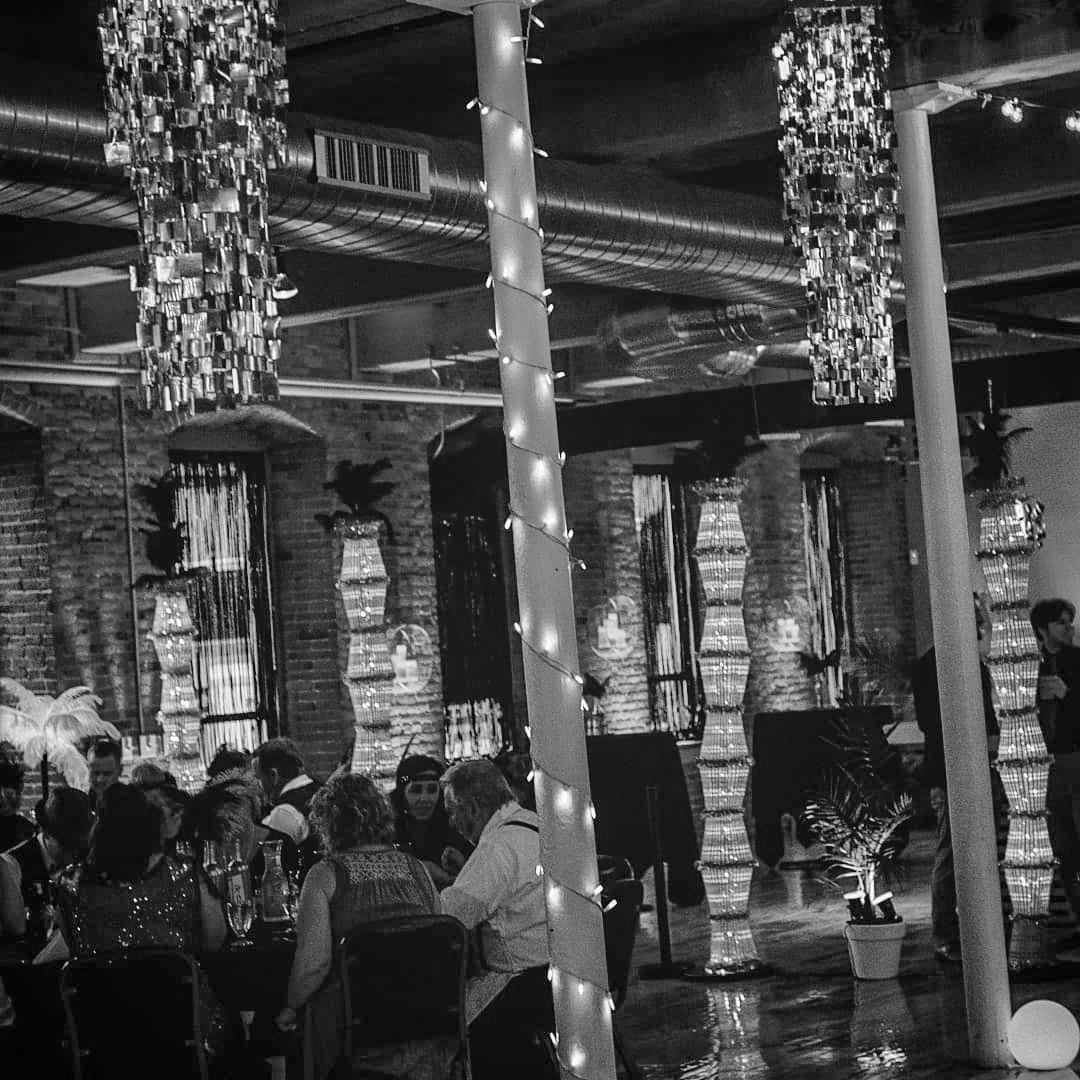 People dining at tables in an industrial-style restaurant with exposed brick walls, large hanging chandeliers, and decorative side lamps in the background. There are string lights wrapped around a central pole and overhead ductwork visible.