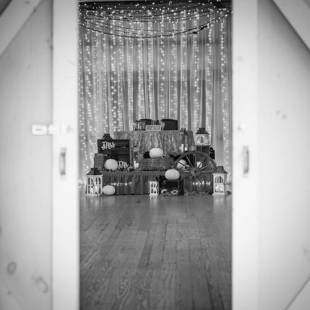 Decorated wedding or event stage with string lights, pumpkins, lanterns, and a wagon wheel, framed by a doorway.