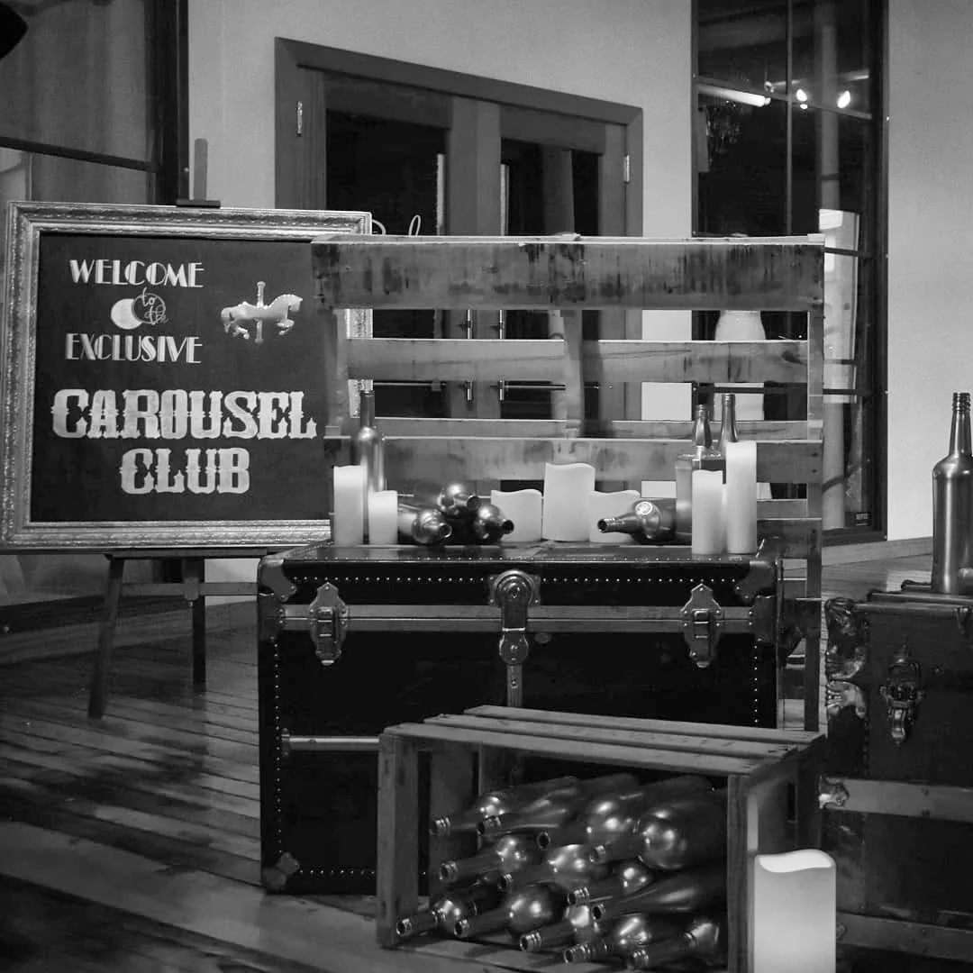 A black and white photo of a sign that says "Welcome to the exclusive Carousel Club" and a wooden display with candles and empty bottles inside. There are additional bottles on a small shelf and a wooden crate filled with more bottles on the floor.