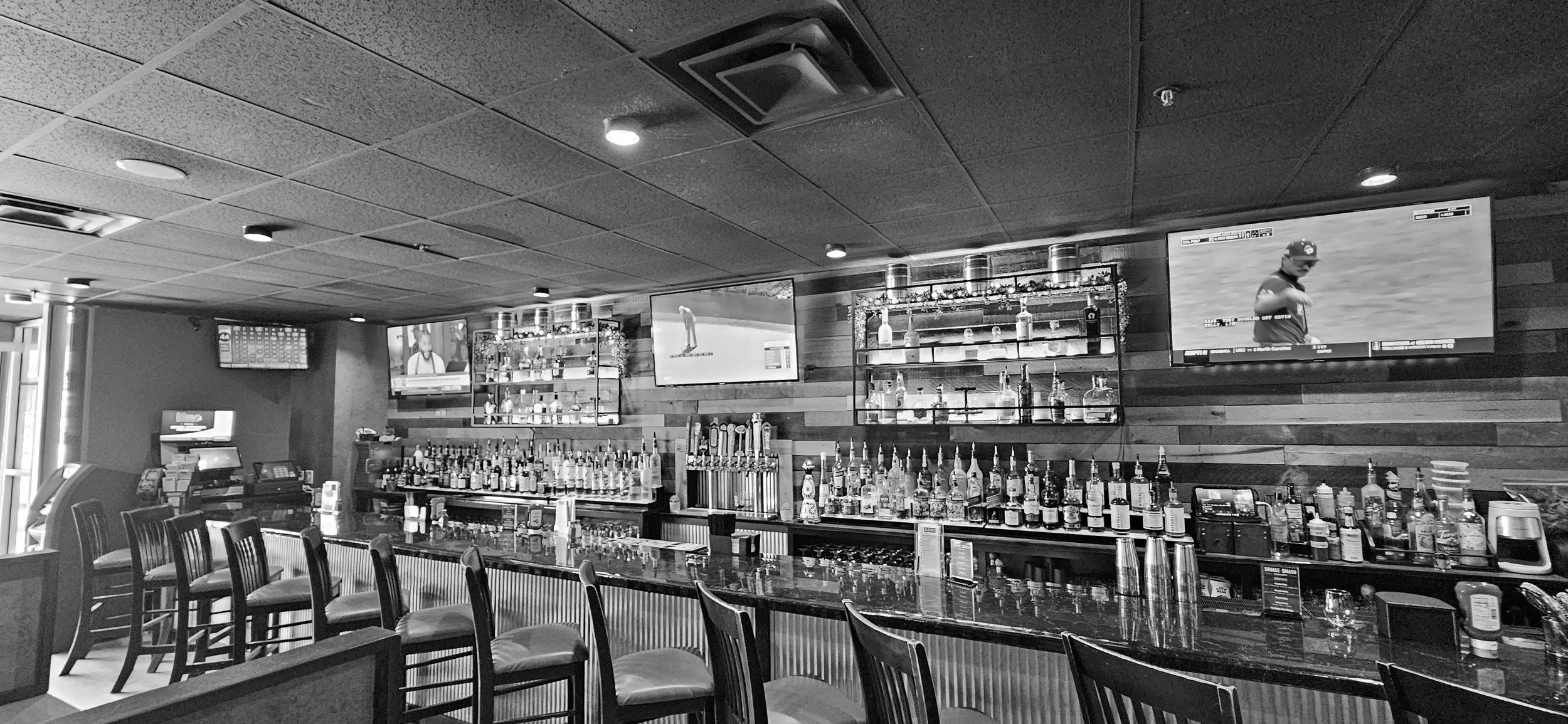 Black and white photo of a bar with liquor bottles on shelves, a long bar counter with chairs, and three television screens displaying sports games on the wall.
