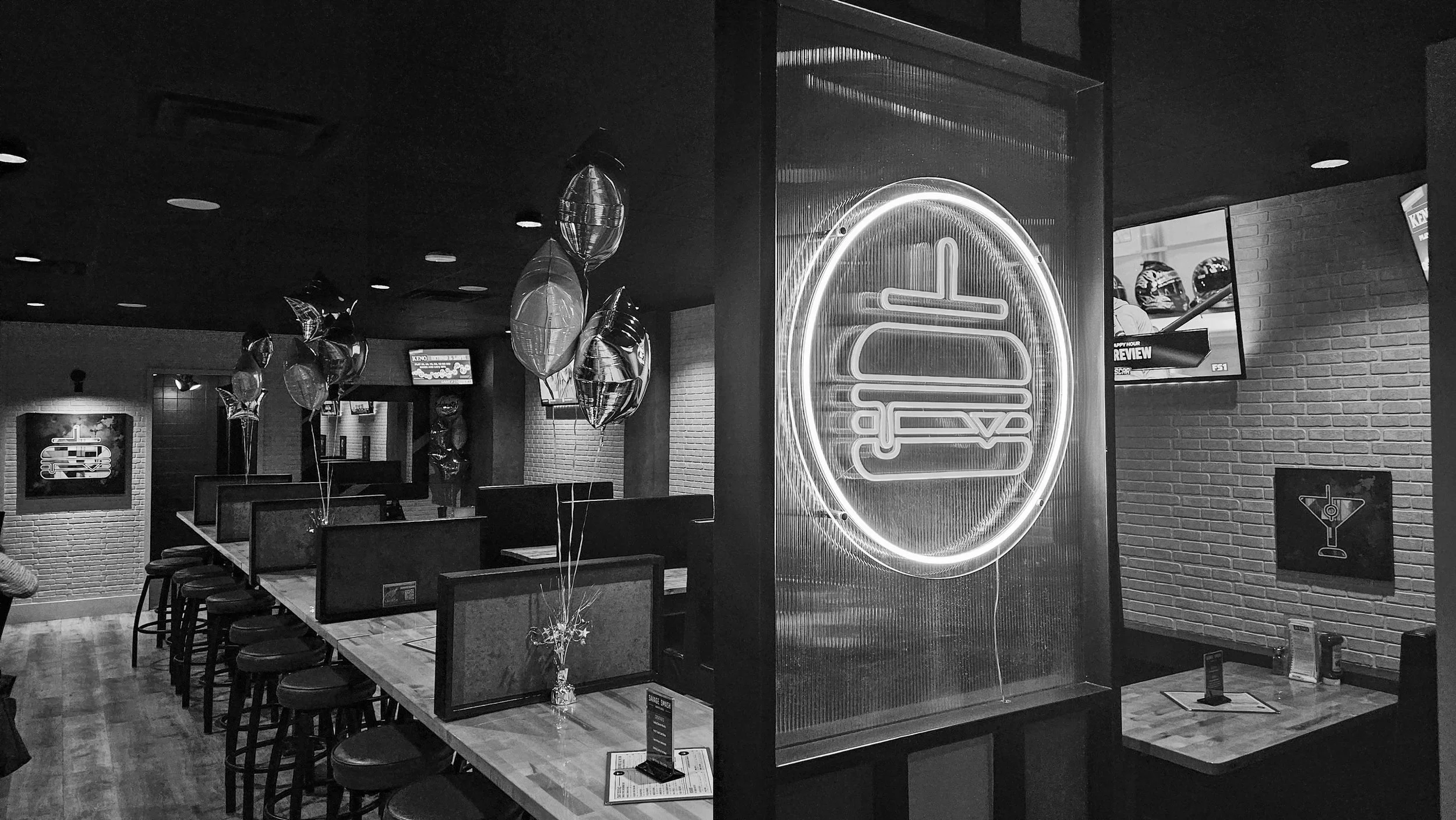Interior of a casual restaurant with tables, chairs, balloons, neon sign, and TV screens, with brick walls and dark ceiling.
