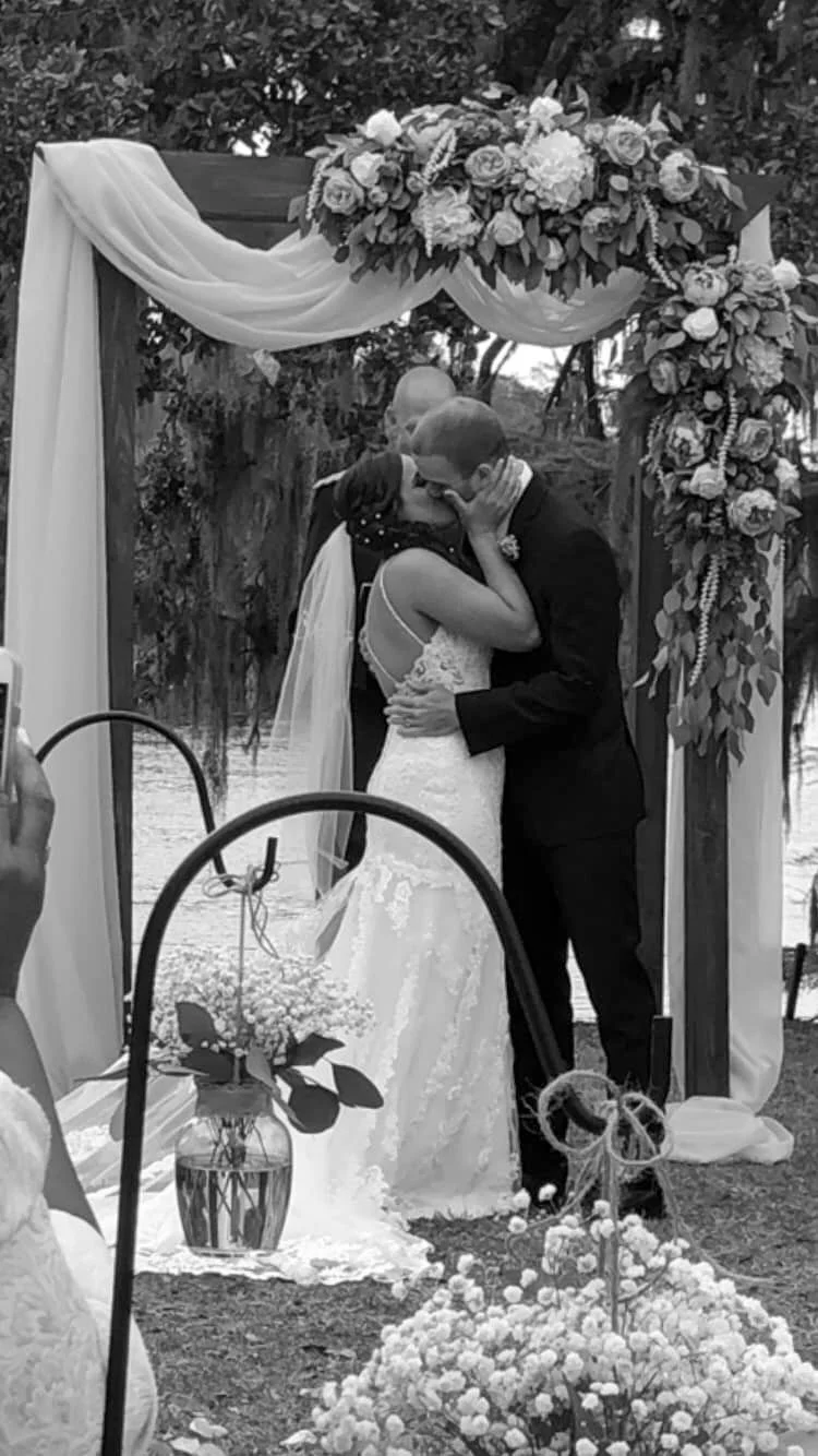 A black and white photo of a wedding ceremony with a bride and groom sharing a kiss under a floral arch.