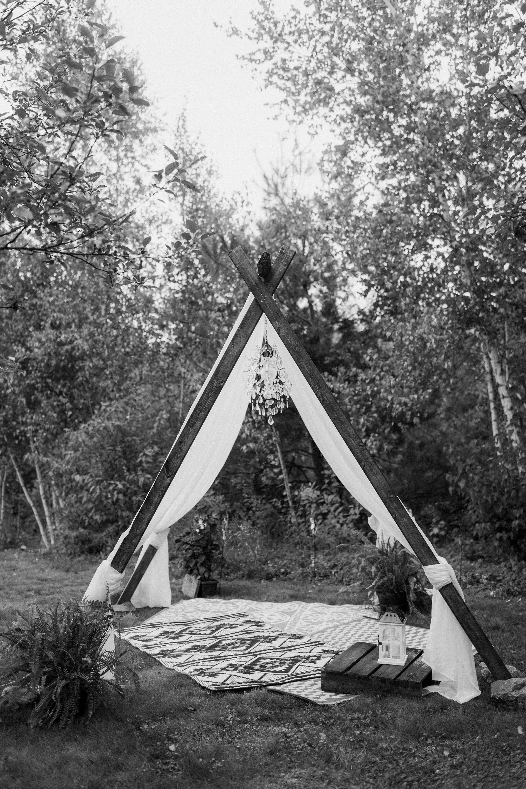 An outdoor wedding setup with an A-frame wooden arch decorated with draped fabric and hanging ornaments. A patterned rug is laid on the grass, and a lantern sits on a small wooden table. The scene is surrounded by trees and foliage.