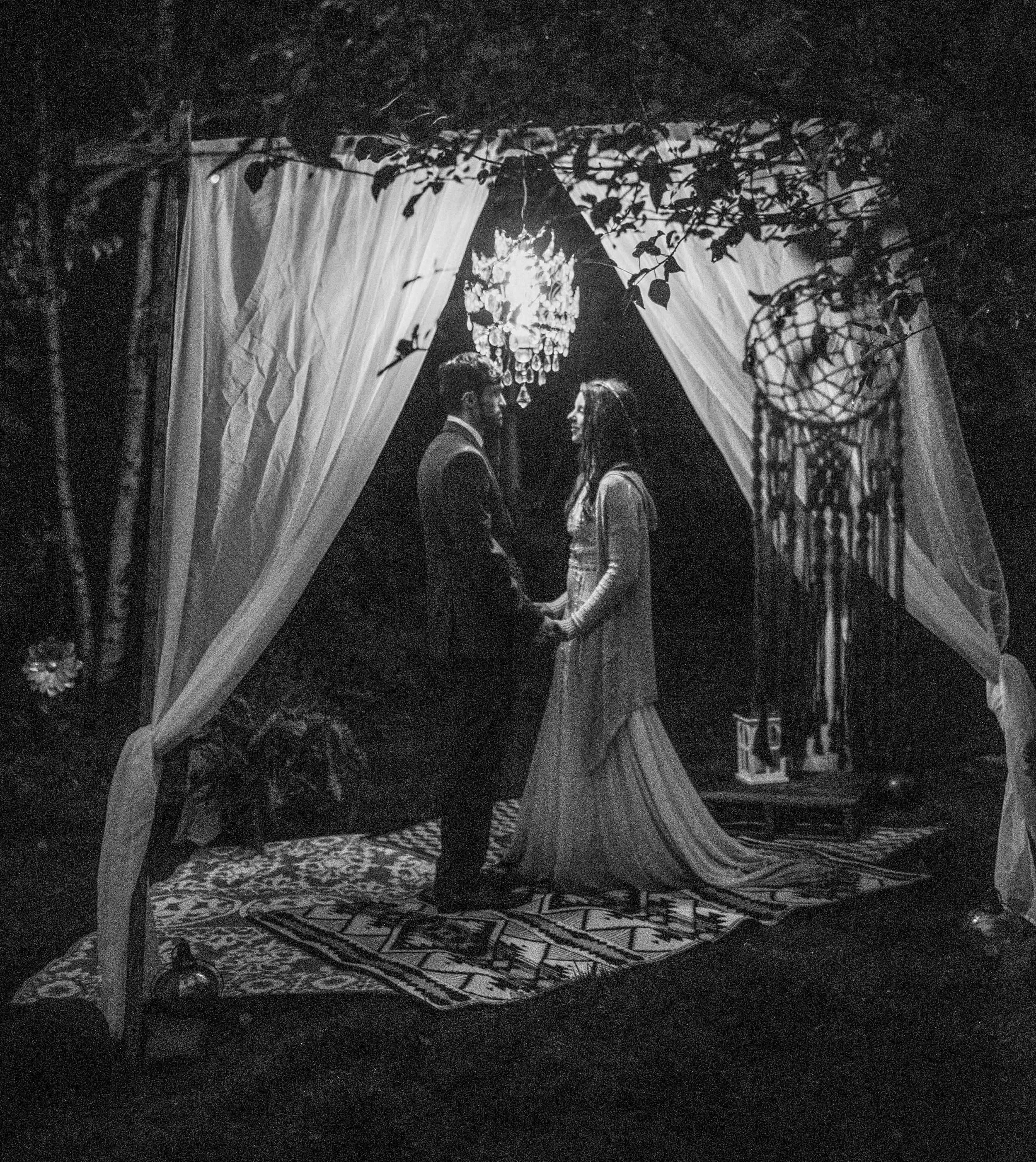 A couple stands facing each other under a decorated canopy with draped curtains, hanging lanterns, and foliage, holding hands and gazing into each other's eyes. The scene appears vintage or artistically styled in black and white.