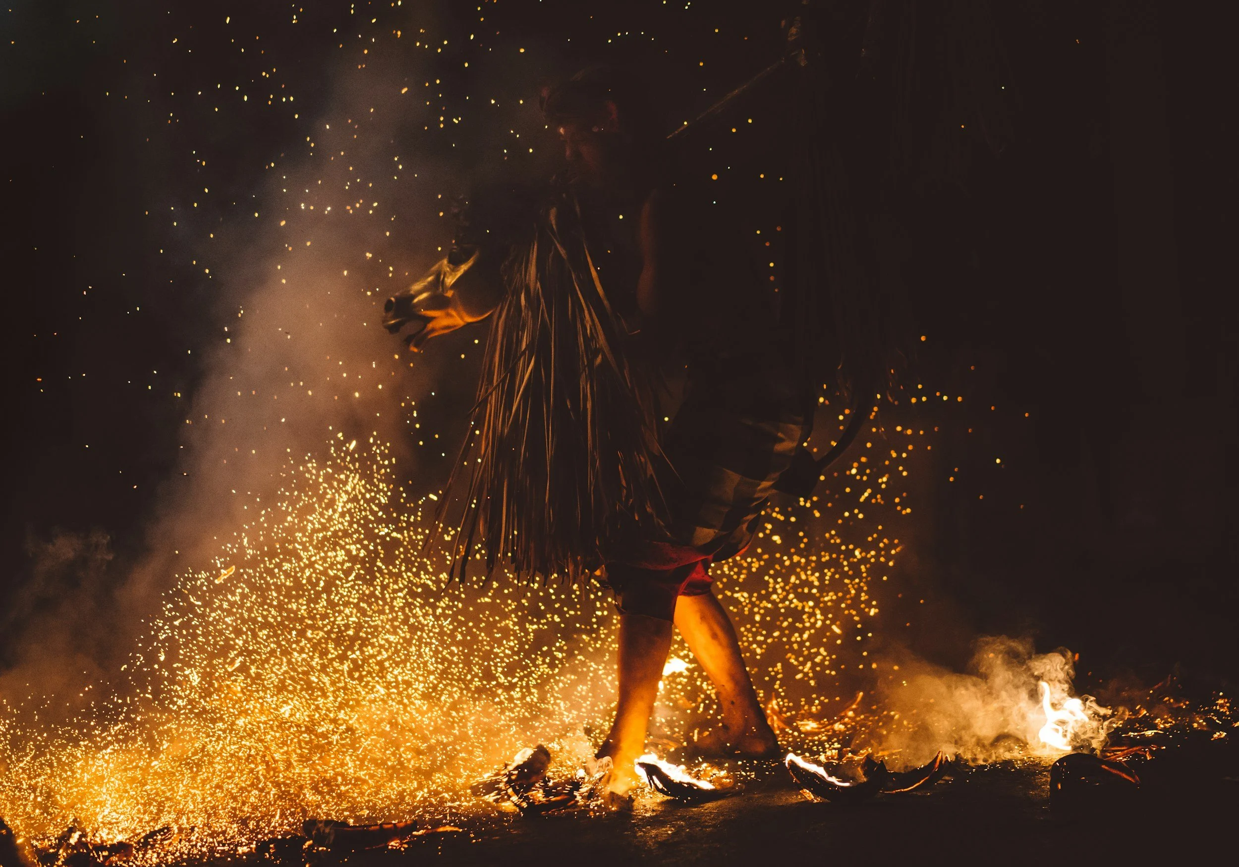 Person participating in a fire performance art, holding a flaming object, surrounded by sparks and smoke at night.