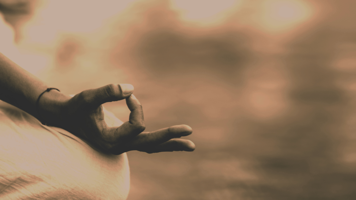 A person sitting cross-legged, making an 'okay' gesture with their hand, during sunset or sunrise over water.