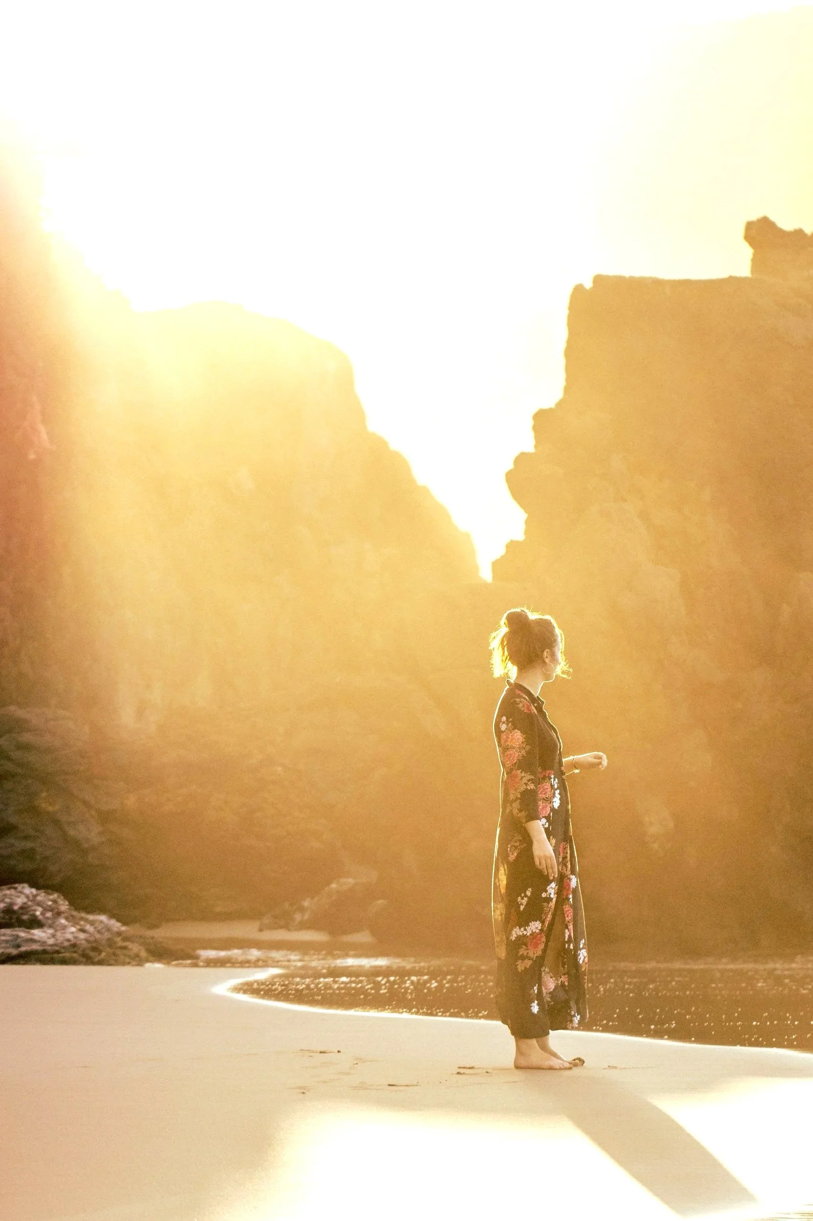 Woman in floral dress walking barefoot on beach at sunset.