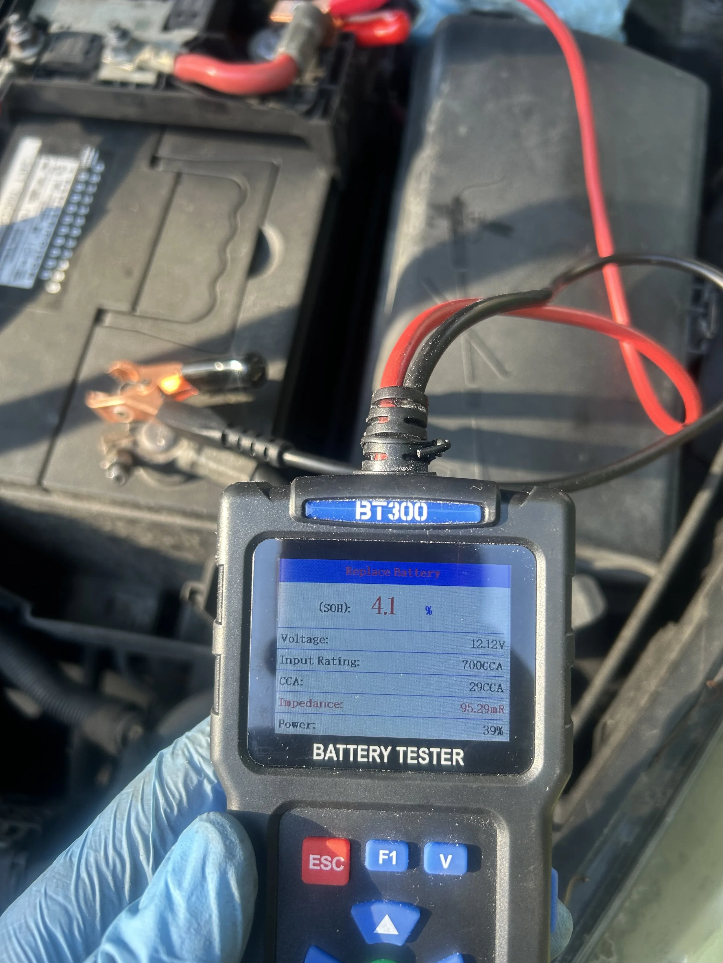 A mechanic wearing a blue glove holding a battery tester in front of a car battery, with red and black cables connected to the battery on a vehicle.