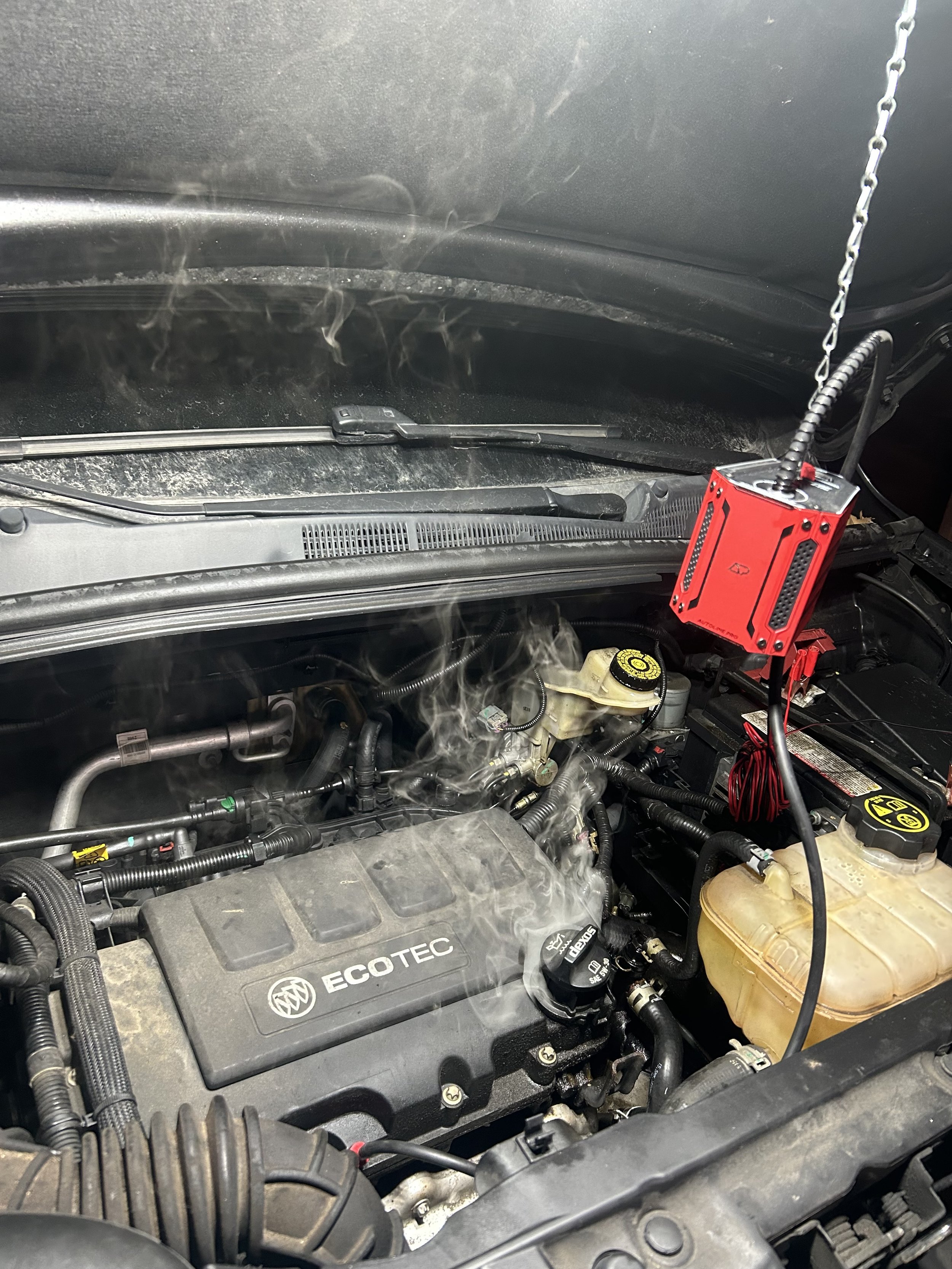 Inside of a car engine bay showing various components including a red diagnostic scanner attached to the engine, a coolant reservoir on the right, and a power steering fluid reservoir in the middle.