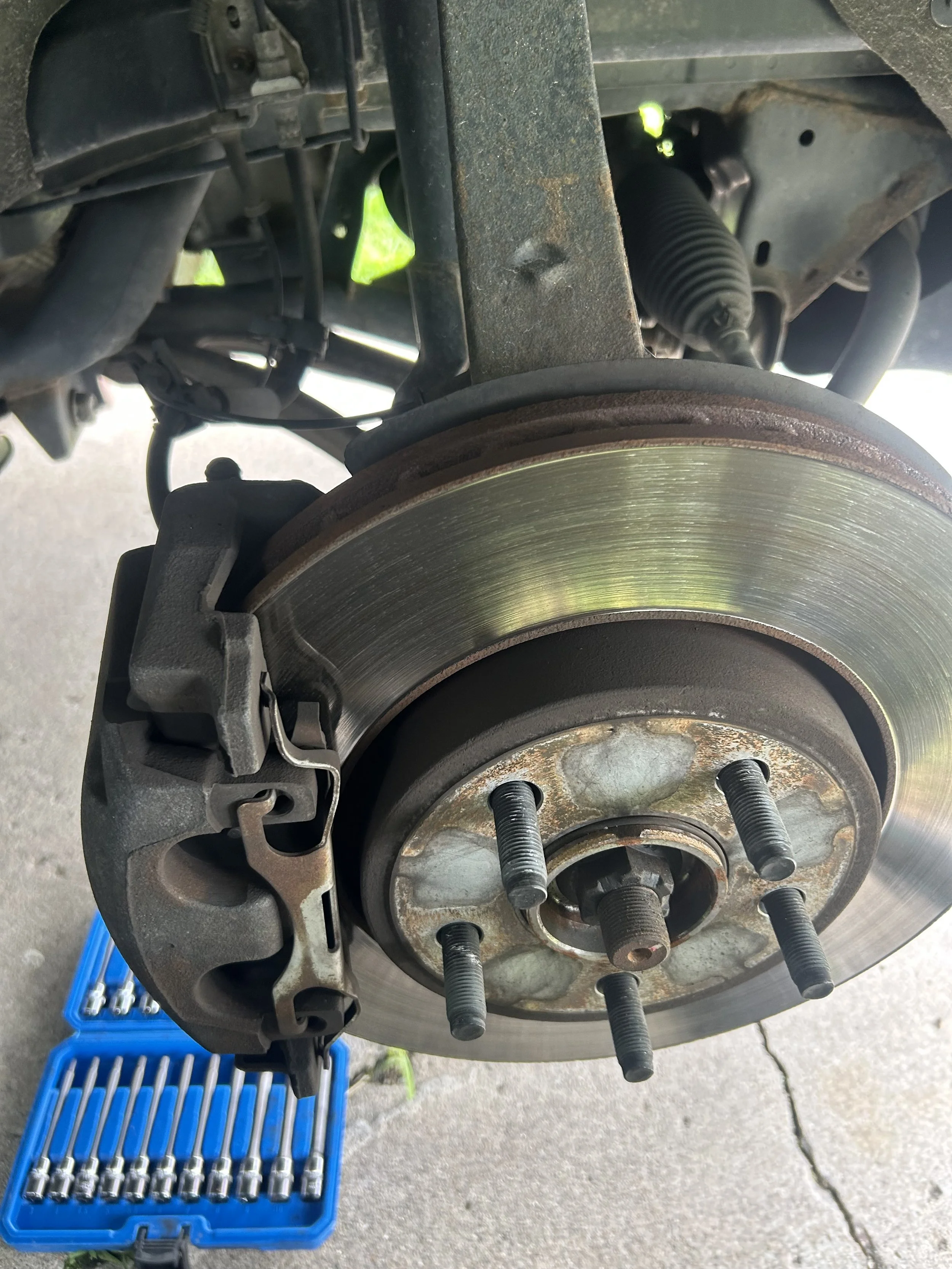 Close-up of a vehicle's wheel hub assembly with a brake rotor, brake caliper, and suspension components visible. A blue toolbox with sockets and ratchets is on the ground nearby.