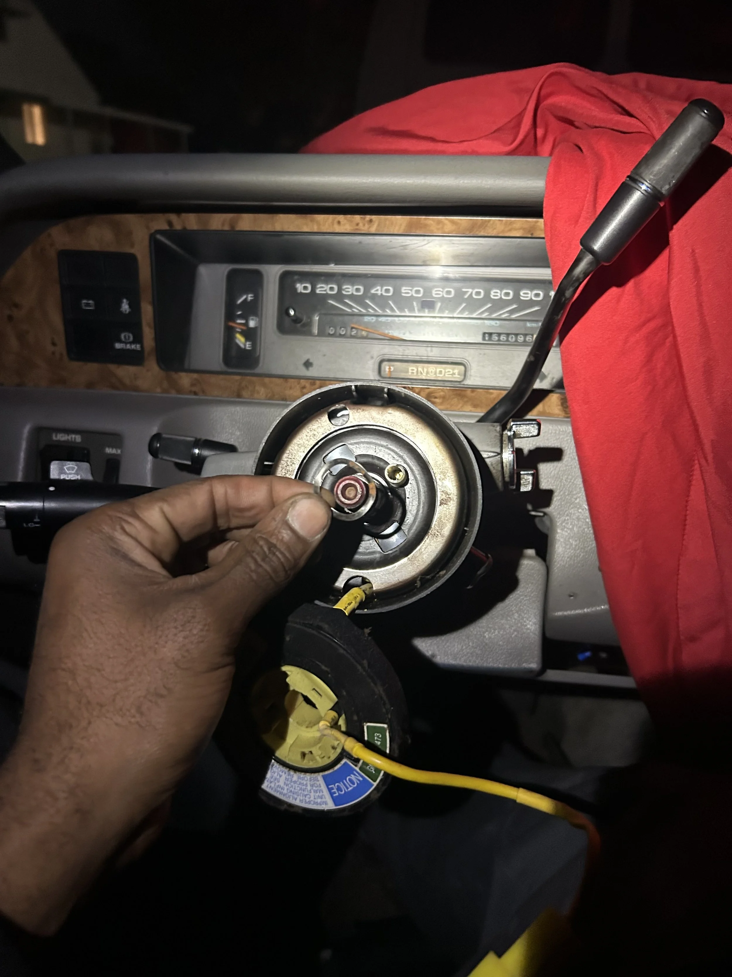 A mechanic working on a car dashboard, holding a steering wheel component with a wiring connector, in a vehicle's interior, with tools and parts visible.