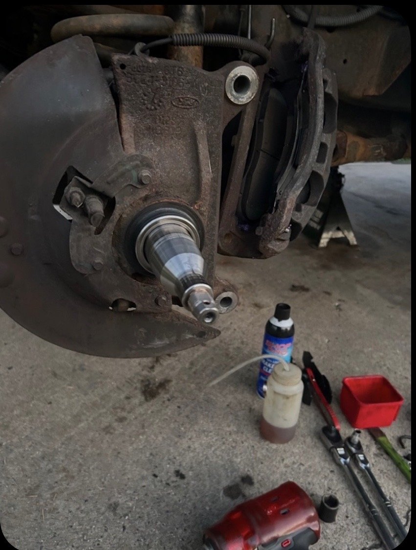 Close-up of an automotive brake assembly, with tools and parts on the ground nearby, including a can of brake cleaner, a container of brake fluid, a wrench, a red tool box, and a power tool.