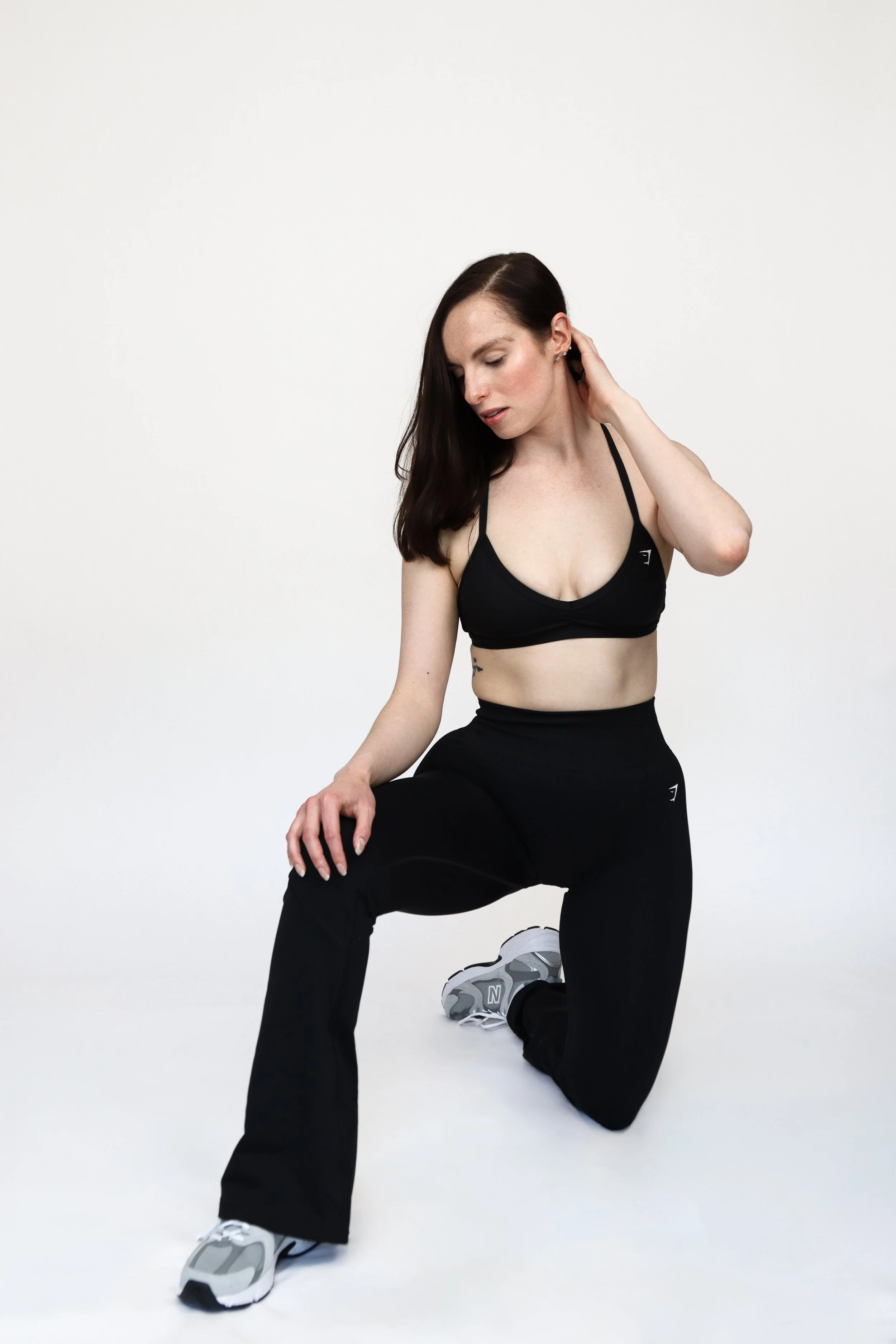 A woman with long dark hair in workout clothes kneeling on one knee and touching her neck in a studio with a white background.
