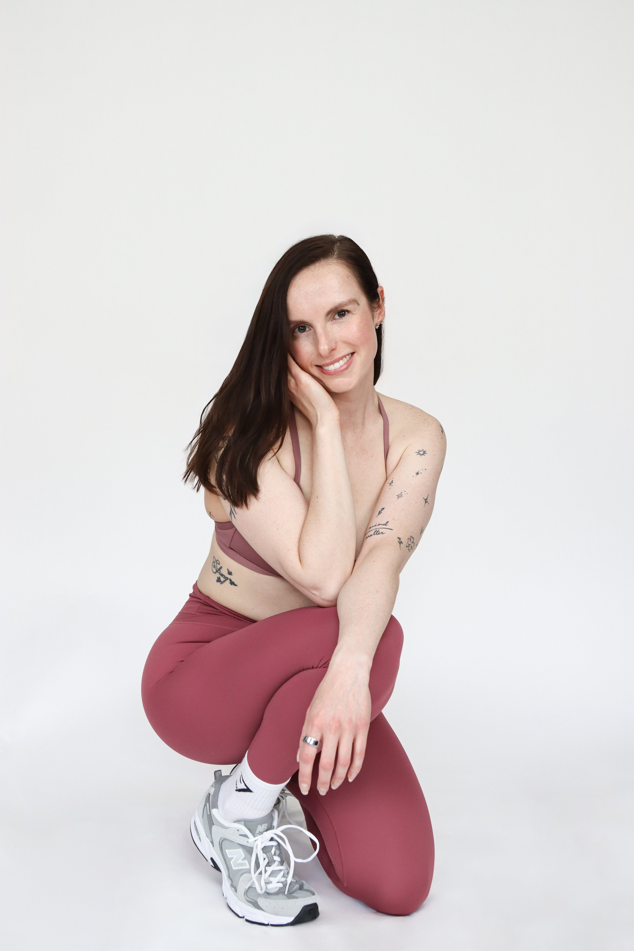 A young woman with long brown hair, tattoos on her arms and stomach, smiling, sitting on one knee and resting her chin on her hand, wearing mauve workout top and matching leggings, white and gray athletic shoes, against a plain white background.