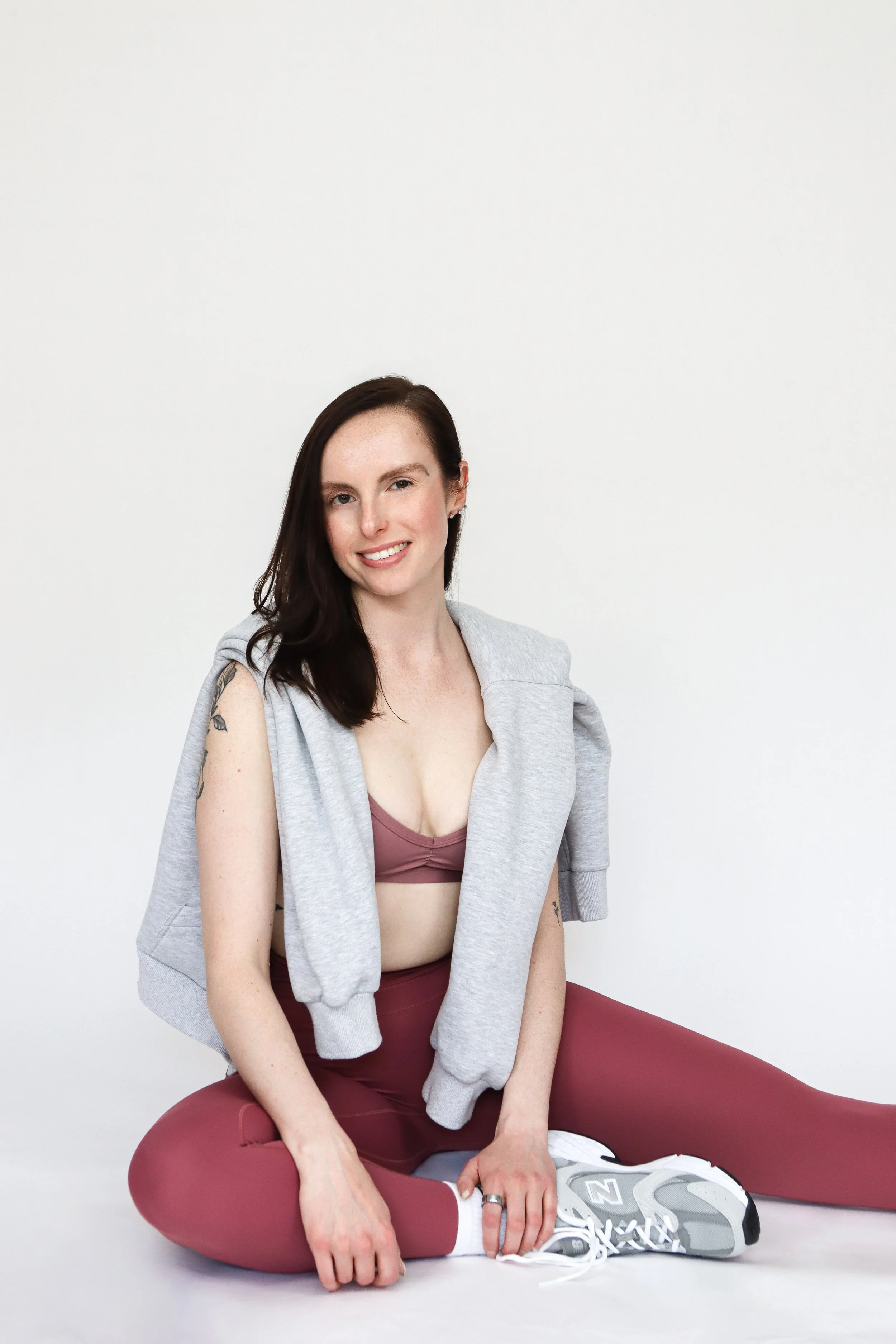 A young woman with dark brown hair, fair skin, and tattoos on her left arm, sitting cross-legged on the floor against a plain white background. She is smiling, wearing a gray sleeveless hoodie, mauve sports bra, matching leggings, and white sneakers. She has a relaxed and confident pose.