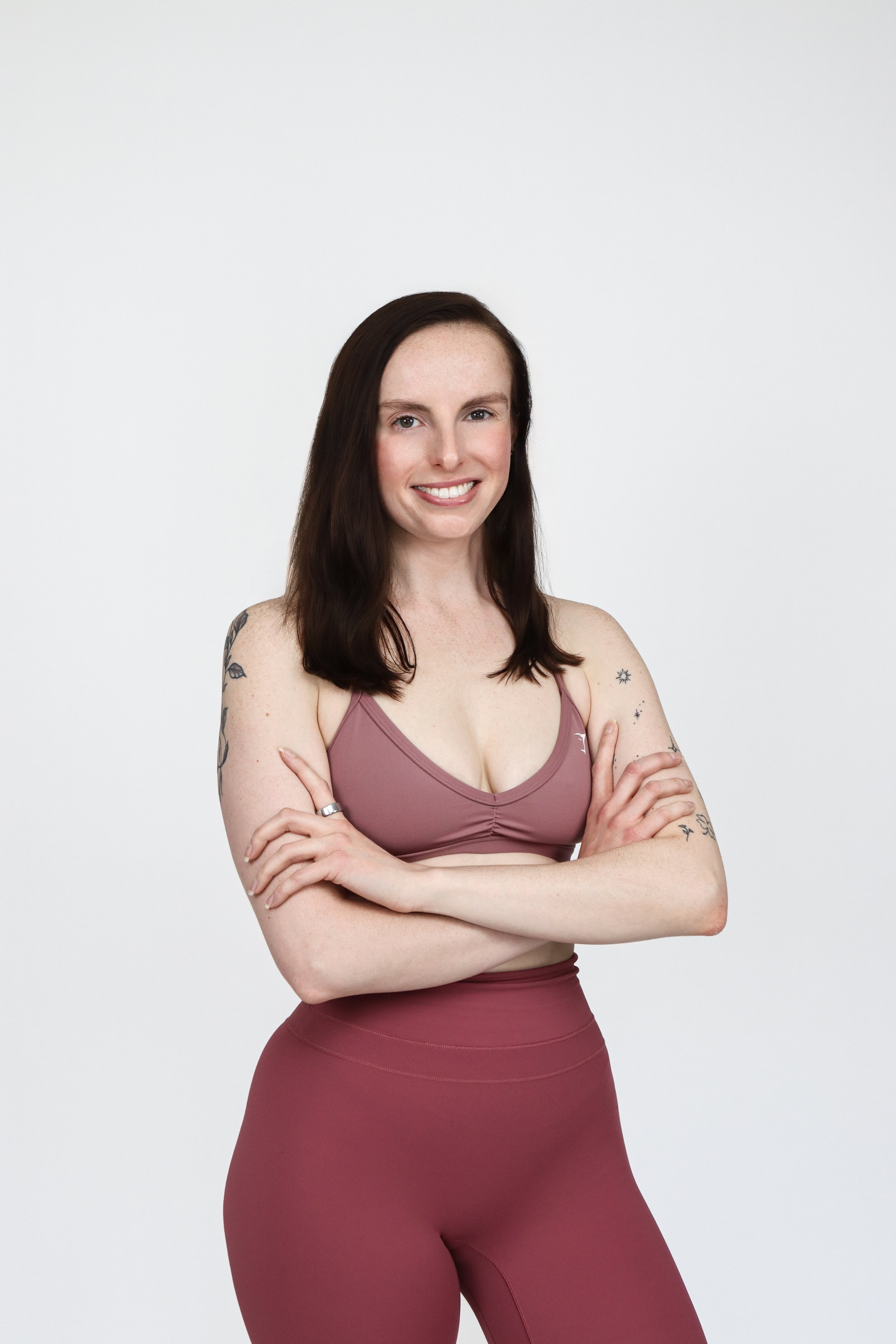 A young woman with dark brown hair and tattoos on her arms, standing with arms crossed and smiling against a white background, wearing a mauve sports bra and matching high-waisted leggings.