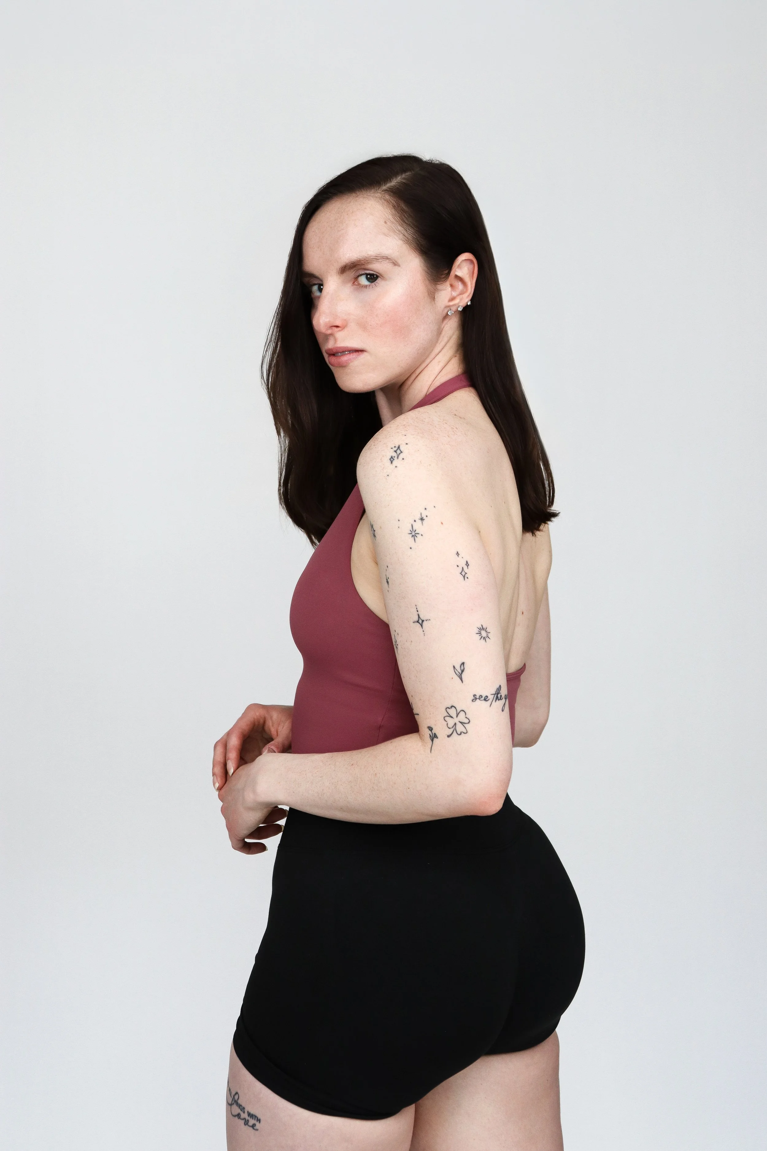 A woman with long dark hair and tattoos on her left arm and thigh, wearing a mauve halter top and black shorts, posing against a plain light gray background.