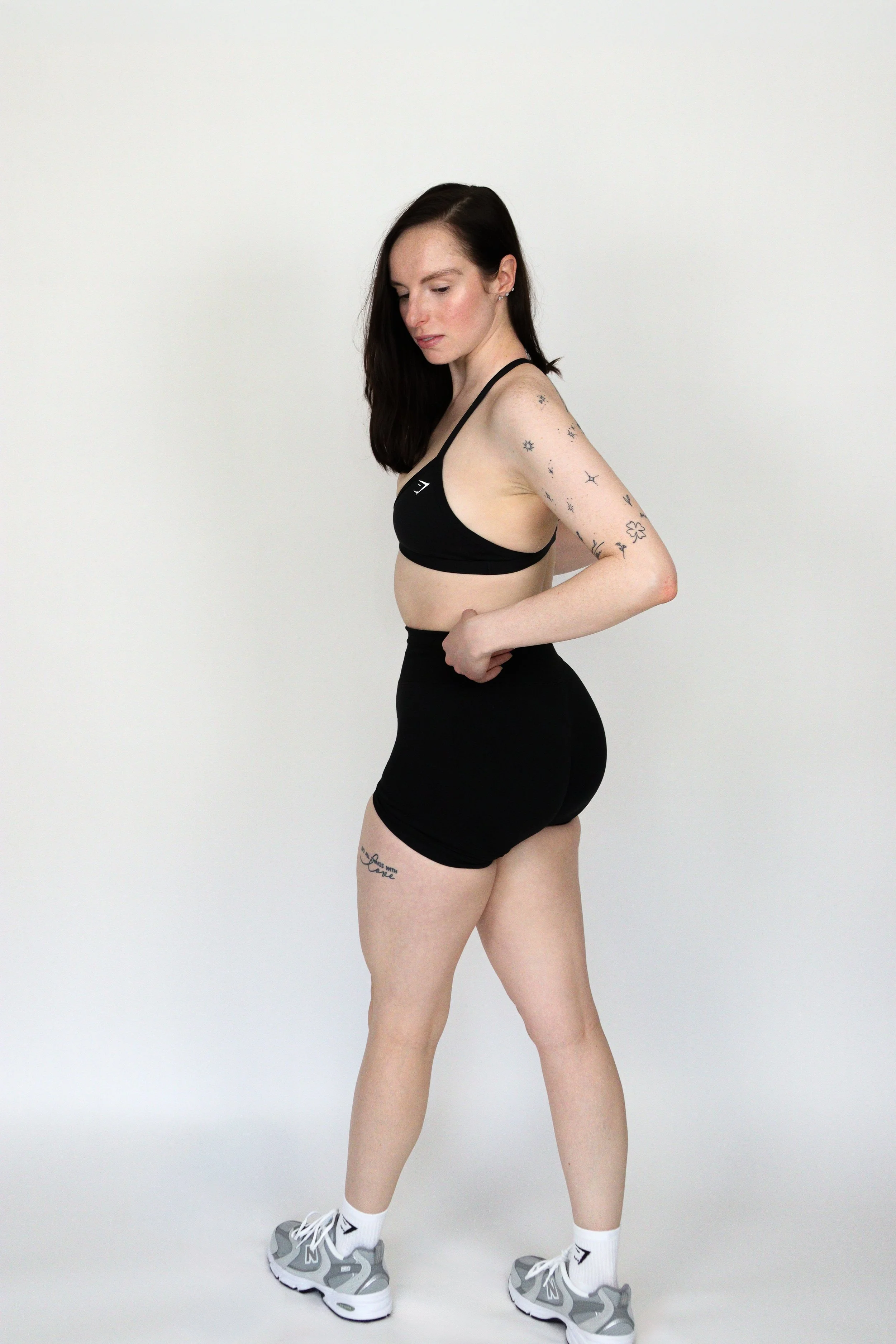 A woman with long dark hair wearing a black sports bra, black shorts, white socks, and gray athletic shoes standing against a plain white background.