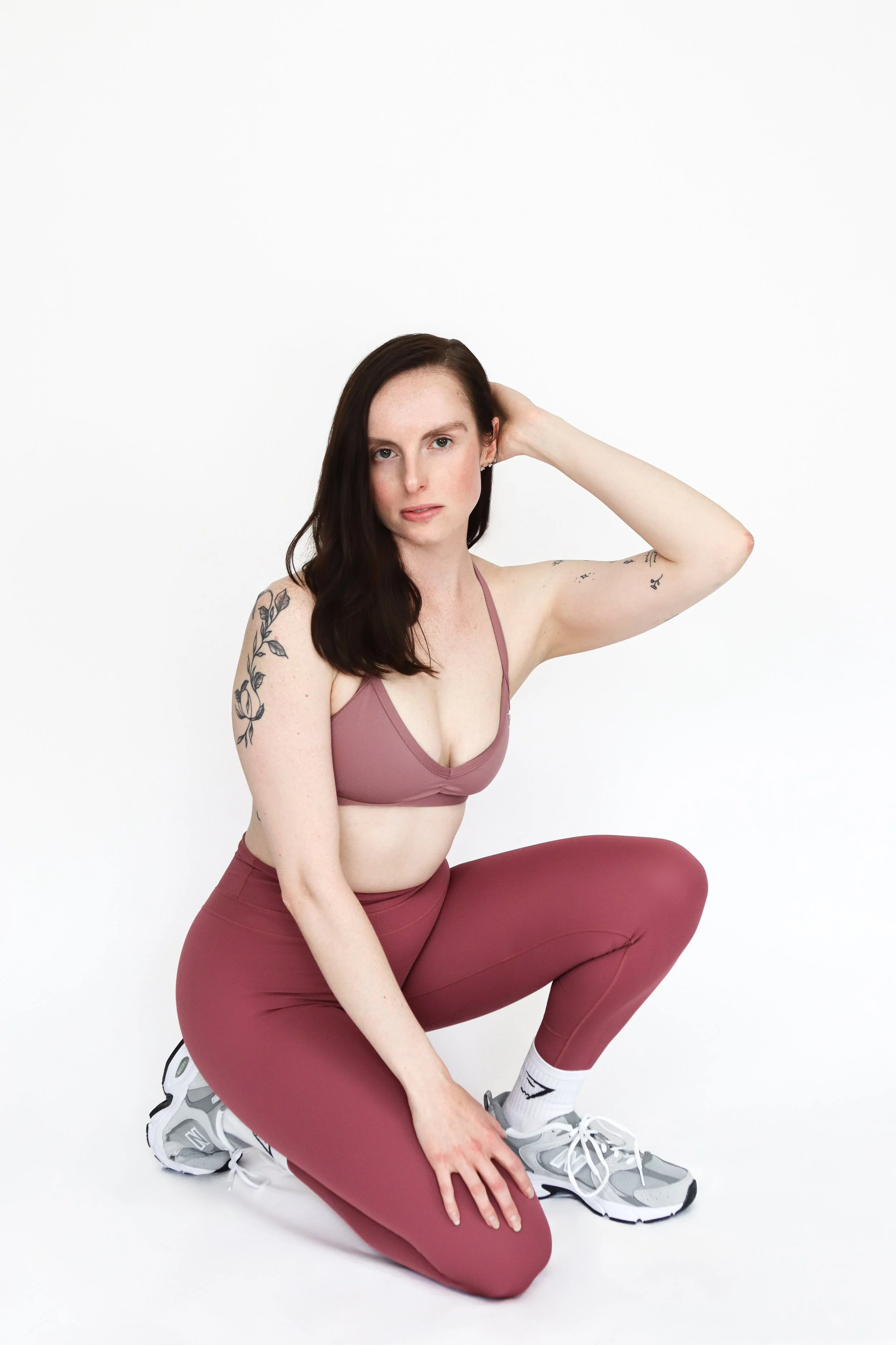 A woman with dark hair, tattoos on her arms, wearing a mauve sports bra and leggings, is in a kneeling pose on a white studio background, with her left hand on her knee and right hand behind her head.