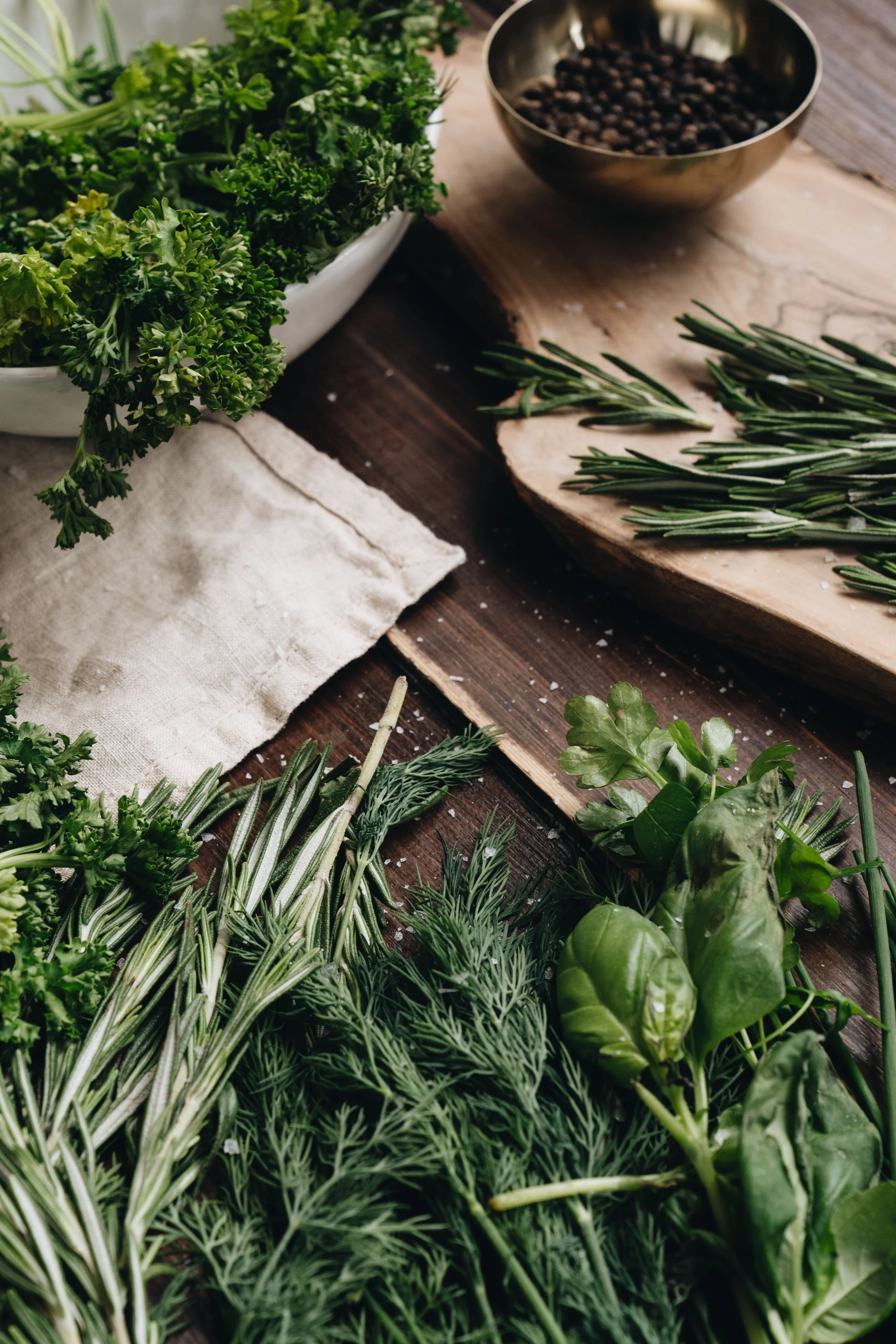 Assorted fresh herbs on a wooden surface, including rosemary, basil, and parsley, with a bowl of black peppercorns nearby.