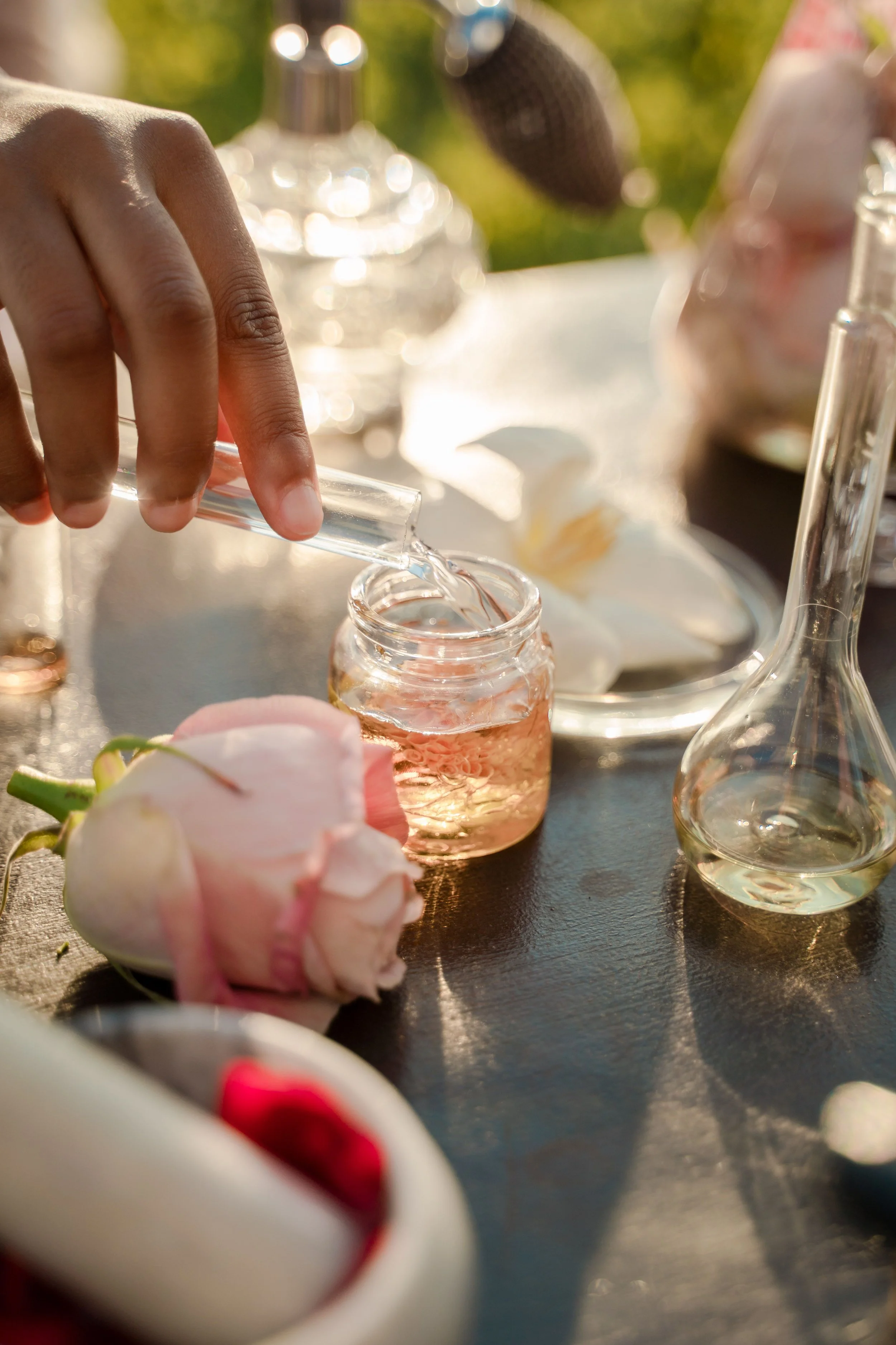 Close-up of a hand pouring a clear liquid from a small test tube into a glass jar with pink liquid inside, with laboratory glassware and blurred flowers in the background, outdoors on a sunny day.