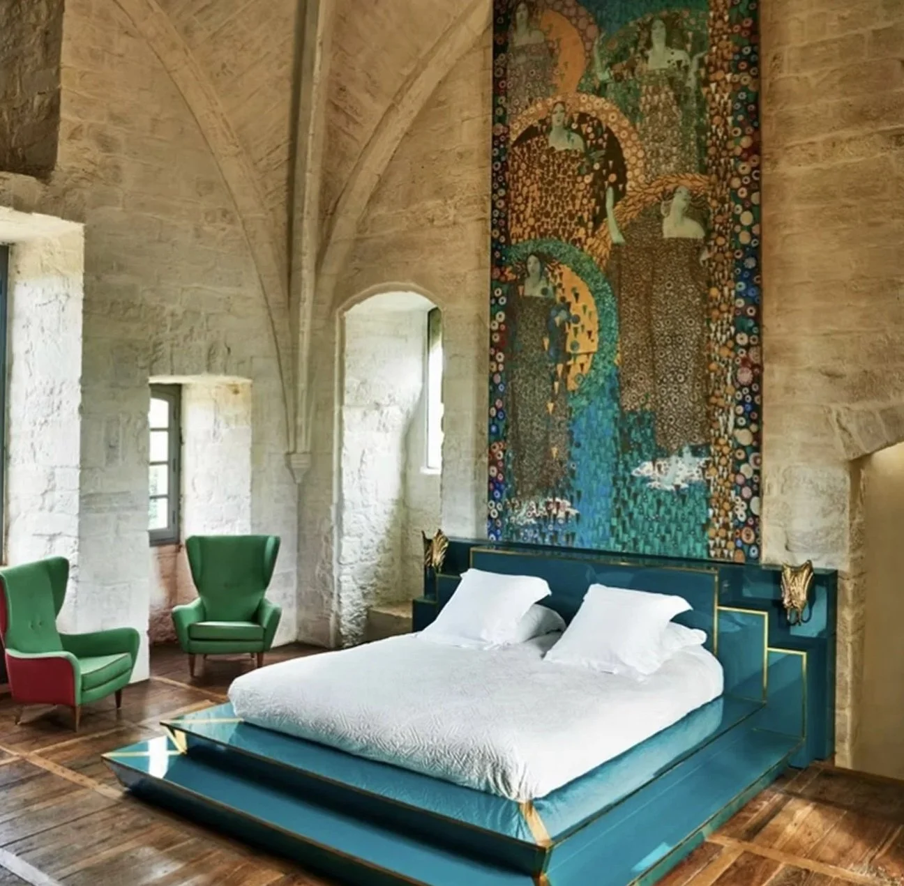 A bedroom with high stone walls and arched ceiling, featuring two windows. The room has two green armchairs and a bed with white linens and a blue and gold headboard. A colorful tapestry hangs above the bed depicting women in patterned clothing.